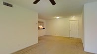 This single-story space features an open-concept living area that serves as the central hub of the unit. The room is finished with wall-to-wall carpeting in a neutral beige tone, contributing to a clean and understated aesthetic. A ceiling fan with dark wood-toned blades is mounted overhead, providing both ventilation and a subtle decorative element.

The living area is illuminated by flush-mount ceiling light fixtures, supplemented by ambient light, giving the space a well-lit atmosphere. White walls throughout keep the interior feeling open and bright. A pass-through window connects the living area to the kitchen, offering a glimpse of cabinetry and countertops within, and allowing light and interaction to flow between the two spaces.

A solid white entry door is positioned along one wall, providing access to the unit and featuring a standard panel design. The overall layout is straightforward and practical, with clean lines and minimal architectural embellishment. The neutral finishes and open floor plan create a versatile living environment that can accommodate a variety of furniture arrangements and personal styles.