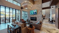 This elegantly designed single-floor communal space blends rustic and contemporary elements to create a warm yet refined atmosphere. The centerpiece of the main lounge is a soaring double-height ceiling accented by dark exposed wood beams, anchored by a striking floor-to-ceiling brick fireplace wall with a mounted flat-screen television above a sleek linear hearth. A large circular chandelier with Edison-style bulbs adds dramatic, ambient lighting to the seating area below, which features plush sofas, leather chairs, and solid wood accent tables arranged for both comfort and conversation. Throughout the space, the herringbone-patterned wood plank flooring adds visual texture and warmth, tying together the various zones seamlessly. Expansive steel-framed, floor-to-ceiling windows and glass doors flood the interior with natural light and provide direct views to an outdoor pool and terrace area. A well-appointed kitchenette features white shaker cabinetry, quartz countertops, a farmhouse sink, subway tile backsplash, exposed brick accents, open wood shelving, and stainless steel appliances — ideal for entertaining. A separate lounge area with additional seating and a game table extends the space further, while curated artwork and accent lighting throughout contribute to the inviting, upscale ambiance.