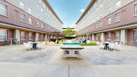 Located at 301 Church Ave in College Station, Texas, this outdoor courtyard is a vibrant communal space enclosed by multi-story brick buildings with a classic architectural aesthetic. The courtyard is thoughtfully designed to offer a variety of recreational and relaxation zones, all centered around a well-maintained turf lawn that serves as the heart of the space.

The hardscaped areas flanking the lawn provide ample room for socializing and activities. Concrete ping-pong tables offer an inviting spot for friendly competition, while overhead shade structures and pergolas create shaded retreats ideal for relaxing. A suspended egg-shaped chair beneath one of the pergolas adds a distinctive decorative touch to the landscaped rock garden nearby.

An outdoor kitchen and bar area, constructed with whitewashed brick and fitted with a built-in grill and dark countertop, is complemented by stylish blue barstools — perfect for casual gatherings. Scattered throughout the courtyard are modern lounge chairs, Adirondack chairs, and bistro table sets that encourage both quiet relaxation and group interaction.

Decorative landscaping, including palm trees, mature shade trees, ornamental boulders, and planted beds, adds texture and greenery throughout. String lights are strung overhead, enhancing the ambiance during evening hours. The brick façades of the surrounding buildings, with their regularly spaced windows, create a sense of enclosure while maintaining an open-sky atmosphere above.