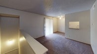 This single-floor unit offers a straightforward and functional layout designed for practical everyday living. The living area is finished with wall-to-wall carpet and features a ceiling fan, a textured ceiling, and a sliding glass door that invites natural light into the space. A half-wall partition separates the living area from adjacent spaces, adding subtle architectural definition. The kitchen is efficiently arranged with warm wood-toned upper and lower cabinetry, a coil-burner electric range, a full-size refrigerator, and a stainless steel sink with a laminate countertop. A range hood overhead and a ceiling vent ensure proper ventilation. Hardwood flooring in the kitchen adds a contrasting warmth to the space. The bathroom is equipped with a full tub-and-shower combination featuring large-format tile surround in earthy tones, a toilet, and a vanity sink. Additional overhead storage cabinets in matching wood tones provide practical organization space. A textured ceiling is consistent throughout the unit, giving the space a cohesive finish. The hallway area, carpeted and simply lit, connects the various spaces efficiently. Overall, the unit presents a classic, no-frills interior with functional amenities suited for comfortable day-to-day living.