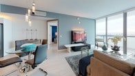 Unit 49B is a thoughtfully designed single-floor residence that seamlessly blends contemporary style with stunning waterfront views. The open-concept living and dining area is anchored by expansive floor-to-ceiling sliding glass doors that frame breathtaking panoramic water views and lead to a private balcony, flooding the space with natural light. The living area features light wood flooring and a soft, neutral color palette accented by cool blue tones throughout.

The kitchen is outfitted with high-gloss white cabinetry, stainless steel appliances including a French door refrigerator and Bosch oven, under-cabinet LED lighting, and a peninsula with bar seating — ideal for both everyday living and entertaining.

The bedroom is a serene retreat, highlighted by a bold blue accent wall, floor-to-ceiling windows with direct water views, and access to the balcony. A spacious walk-in closet adjacent to the bedroom offers custom built-in shelving, drawers, and hanging space, with light wood flooring connecting through to the en-suite bathroom featuring marble-look tile flooring and clean white finishes.

A secondary living space with a textured concrete feature wall and unique wall-mounted bicycle display adds an urban, industrial character. The unit also includes a powder room with a sleek undermount sink and frameless mirror.