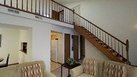 This two-story residence makes a strong first impression with its dramatic double-height ceiling in the main living area, which creates an open, airy atmosphere throughout the home. The living room is a welcoming space, furnished with comfortable upholstered armchairs accented with patterned cushions, centered around a compact accent table — a perfect setup for relaxed gatherings or quiet evenings.

A prominent staircase with rich, dark wood banisters and spindles rises gracefully from the main floor to the upper level, serving as both a functional and aesthetic centerpiece of the home. The upper floor features an open railing overlooking the living area below, reinforcing the sense of vertical space and connectivity between floors.

The kitchen is visible from the living room and offers a functional layout with standard appliances, including a full-size refrigerator, and cabinetry that provides ample storage. The transition between the living area and kitchen is seamless, lending to an open-concept feel on the main level.

Additional doors visible on the main floor suggest access to closets, utility spaces, or further rooms, adding practical storage and versatility to the layout. The neutral color palette throughout — soft whites and warm beiges — gives the space a clean, timeless look that is easy to personalize.