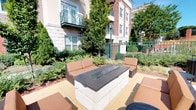 This thoughtfully designed space seamlessly blends outdoor and indoor living. The centerpiece of the outdoor area is a built-in fire pit with a sleek stone base and dark granite top, surrounded by comfortable cushioned seating arranged to encourage gathering and conversation. Lush, manicured greenery, mature trees, and well-maintained garden beds frame the lounge area, while an ornamental iron fence and classic brick building facades provide an elegant urban backdrop.

Inside, the interior rooms are finished with warm, neutral tones throughout. Plush carpeted floors extend across the bedroom spaces, each benefiting from natural light streaming through blinded windows that offer pleasant outdoor views of green surroundings. The rooms feature clean white trim and baseboards, creating a crisp contrast against the soft beige walls.

A well-equipped bathroom is accessible from the bedroom area, showcasing rich dark wood cabinetry, a granite-topped vanity with a large mirror, hardwood-style flooring, and a full bathtub and shower combination. The kitchen, visible through a connecting doorway, features pendant lighting, dark cabinetry, a granite island, and the same warm hardwood-style flooring, tying the interior aesthetic together cohesively. Overall, the space offers a comfortable, polished environment suited for everyday living.