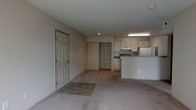 Located at 6785 Oakton Ln in Columbus, Ohio, this single-floor residence offers a clean and functional layout well-suited for everyday living. The main living area is carpeted and open, providing ample room to arrange furnishings freely. Natural light enters through the front entrance, which features a classic six-panel door with a tiled entryway mat area, creating a defined transition from the outdoors. The space flows seamlessly into a compact kitchen, which is separated from the living area by a raised countertop bar that doubles as a casual dining or workspace surface. The kitchen is equipped with white cabinetry offering generous storage, a range with overhead ventilation, a refrigerator, and under-counter appliances — all set against neutral-toned walls and flooring. A closet with sliding doors is positioned adjacent to the kitchen area, providing convenient built-in storage. The overall color palette throughout the space is neutral, with beige walls and light carpeting that create a calm, understated ambiance. The layout is straightforward and practical, making efficient use of the available space while maintaining an open and airy feel throughout.
