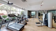 This fitness center offers a comprehensive workout environment thoughtfully designed for both cardio and strength training. The space is finished with light hardwood-style flooring that runs throughout, lending a clean and modern aesthetic underfoot. Large floor-to-ceiling windows along one wall flood the interior with natural light and provide views of surrounding greenery, creating an energizing atmosphere during workouts.

The cardio zone features multiple treadmills, elliptical trainers, and a stationary bike, all positioned to take advantage of the natural light and outdoor views. The strength-training area is equally well-appointed, including a multi-functional cable machine, a weight rack with dumbbells, a Smith machine or squat rack, adjustable benches, and functional training accessories such as medicine balls and resistance tools.

A dedicated stretching and floor exercise zone is defined by padded rubber flooring and is anchored by a wall lined with three large framed mirrors, which visually expand the space and allow users to monitor their form. The surrounding walls display a textured, woven-style wall treatment that adds a refined design element. Recessed lighting throughout ensures consistent illumination, complementing the natural light and contributing to the space's polished, welcoming ambiance.
