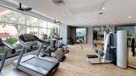 This fitness center offers a comprehensive workout environment thoughtfully designed for both cardio and strength training. The space is finished with light hardwood-style flooring that runs throughout, lending a clean and modern aesthetic underfoot. Large floor-to-ceiling windows along one wall flood the interior with natural light and provide views of surrounding greenery, creating an energizing atmosphere during workouts.

The cardio zone features multiple treadmills, elliptical trainers, and a stationary bike, all positioned to take advantage of the natural light and outdoor views. The strength-training area is equally well-appointed, including a multi-functional cable machine, a weight rack with dumbbells, a Smith machine or squat rack, adjustable benches, and functional training accessories such as medicine balls and resistance tools.

A dedicated stretching and floor exercise zone is defined by padded rubber flooring and is anchored by a wall lined with three large framed mirrors, which visually expand the space and allow users to monitor their form. The surrounding walls display a textured, woven-style wall treatment that adds a refined design element. Recessed lighting throughout ensures consistent illumination, complementing the natural light and contributing to the space's polished, welcoming ambiance.