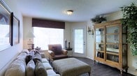 This single-floor living space offers a welcoming and well-appointed interior centered around a comfortable living area. Dark hardwood floors run throughout, providing a rich contrast to the light-toned walls and textured popcorn ceiling overhead. A flush-mount ceiling fixture provides ambient lighting, complementing the natural light that flows generously through a large window dressed with a deep burgundy valance and an adjacent glass-panel exterior door. The living area is furnished with a plush sectional sofa accented with animal-print cushions, a matching leopard-print ottoman that doubles as a coffee table, and a coordinating upholstered armchair. A wooden side table and decorative accessories, including sculptural accents and wall-mounted iron sconces, add character to the space. A prominent natural wood display cabinet with glass panels stands along one wall, offering both storage and a showcase for decorative items and lush potted greenery. The overall ambiance is warm and inviting, blending eclectic furnishings with classic touches to create a lived-in, personalized atmosphere.