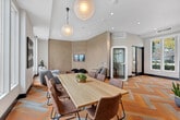 Located at 2225 Collett Ave in Corona, California, this single-floor community space offers a thoughtfully designed open-plan layout combining a dining area and a living room into one cohesive, welcoming environment. The most immediate standout feature is the eye-catching herringbone-patterned flooring in warm amber and neutral tones, which anchors the entire space with bold visual energy.

Large, floor-to-ceiling windows line the exterior walls, flooding the interior with generous natural light and offering direct views of an outdoor pool and patio area. The dining zone features a sleek light wood table surrounded by leather-upholstered chairs, centered beneath a sculptural globe pendant light. Curated artwork adorns the walls throughout, adding an artistic, residential warmth to the space.

The adjacent living area provides comfortable lounge seating, including a soft gray sectional and accent chairs, arranged atop a layered area rug. A textured accent wall adds depth and contrast, while recessed lighting and additional pendant fixtures ensure well-distributed illumination. A private pod or phone booth enclosure and a built-in counter area with warm wood cabinetry offer functional versatility. The overall ambiance is modern and inviting, blending contemporary design with a relaxed, community-focused atmosphere.