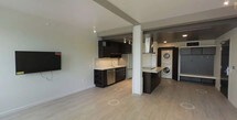 This single-floor space showcases a contemporary open-concept layout that seamlessly connects the living area and kitchen, creating a functional and visually cohesive environment. The kitchen is outfitted with dark, floor-to-ceiling cabinetry that provides generous storage, complemented by light-toned countertops that offer a striking contrast. Stainless steel appliances, including a refrigerator and dishwasher, add a polished, modern finish, while a wall-mounted range hood anchors the cooking area. Track lighting runs along the ceiling, providing focused illumination throughout the kitchen space. A structural column serves as a subtle architectural divider between the kitchen and an adjacent area that houses a stacked washer and dryer unit — a highly practical in-unit convenience. The light wood-toned flooring flows continuously throughout, lending warmth and cohesion to the overall aesthetic. Neutral gray walls keep the palette clean and versatile, while the open layout enhances the sense of spaciousness. The design balances modern industrial elements with everyday functionality, making it a well-appointed and efficient living space.