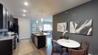 This 2-bedroom, 2-bathroom residence offers a thoughtfully designed single-floor layout that maximizes both functionality and style. The open-concept living area seamlessly connects the kitchen, dining, and living spaces, creating an inviting flow throughout the home. The kitchen serves as a central hub, anchored by a large island with a granite countertop, undermount sink, and pendant lighting overhead. Dark cabinetry provides a bold contrast against the light countertops, while stainless steel appliances complete the modern aesthetic. A dedicated dining area sits adjacent to the kitchen, defined by a charcoal accent wall that adds depth and visual interest to the space. Rich hardwood-style flooring runs throughout the main living areas, complementing the neutral color palette accented by soft teal and gray tones on the walls. The living room, visible from the kitchen, benefits from natural light streaming through large windows fitted with roller shades. Recessed lighting throughout ensures the space is well-lit at all times. The overall ambiance is contemporary and polished, offering a comfortable and stylish environment well-suited for both everyday living and entertaining.