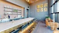 Located at 4126 River Rd in Wilmington, NC, this upscale rooftop amenity space combines a refined interior clubroom with a generously sized outdoor terrace, offering an elevated social environment with sweeping views of the surrounding landscape and waterway.

The interior is anchored by a dramatic floor-to-ceiling curtain wall that floods the space with natural light and frames panoramic outdoor views. Soaring ceilings with a distinctive geometric copper-toned canopy add architectural depth and warmth. A central island with a white quartz countertop, fluted wood base, and gold-accented bar seating serves as the social focal point. A full back bar features open glass shelving, an under-counter refrigerator, and a warm tile backdrop illuminated by integrated lighting. Decorative gold wave art panels accent the muted gray walls, lending a refined, contemporary aesthetic. Large-format tile flooring extends throughout, connecting the interior seamlessly to the transitional hallway areas.

The expansive outdoor terrace wraps around the building's exterior, offering multiple distinct seating zones furnished with weather-resistant sofas, lounge chairs, dining sets, and curved sectionals. Lush planters with manicured greenery define the space, while open railings preserve unobstructed views of the treetops and river beyond — ideal for both casual gatherings and curated events.