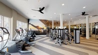 The Fitness Studio at 1670 Metro in Chula Vista is a spacious, single-floor facility thoughtfully designed to support a comprehensive workout experience. The studio features a bold chevron-patterned carpet in neutral tones of beige and gray that runs throughout, creating a dynamic visual foundation while providing a comfortable surface underfoot.

The cardio zone is lined with Life Fitness treadmills positioned along a wall of floor-to-ceiling windows, flooding the area with natural light and offering views of the surrounding outdoor environment. Additional cardio equipment, including elliptical machines and steppers, is arranged along one side of the studio, complemented by a striking large-scale motivational mural depicting an athlete silhouetted against a dramatic sunset sky.

The strength training area is generously outfitted with a full rack of dumbbells, adjustable benches, and multiple selectorized weight machines catering to a wide range of muscle groups. Full-length mirrors line the walls, amplifying the sense of space and allowing users to monitor their form. Structural columns are integrated throughout, adding an architectural rhythm to the open floor plan.

A dedicated amenity station includes a mounted flat-screen TV, a storage credenza, a water station, and sanitizing supplies. Ceiling fans and recessed lighting throughout ensure a well-ventilated and well-lit environment conducive to focused, effective training.