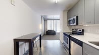 This single-floor unit showcases a distinctive industrial-modern aesthetic defined by exposed concrete structural columns, concrete ceiling soffits, and polished concrete floors that flow through the main living and kitchen areas. The open layout connects the kitchen directly to the living space, creating an efficient and cohesive floor plan.

The kitchen is well-equipped with a full suite of stainless steel appliances, including an electric range, over-the-range microwave, dishwasher, and refrigerator. Gray cabinetry lines the upper and lower walls, complemented by a white countertop and an undermount sink. A compact dining area with a dark wood table and bar-style seating sits adjacent to the kitchen.

The living area transitions to carpeted flooring and features large floor-to-ceiling windows fitted with horizontal blinds, allowing generous natural light to filter into the space. Exposed concrete columns and ceiling details reinforce the industrial character throughout.

The bathroom offers a clean, functional design with small-format mosaic tile flooring, a wall-mounted vanity with a mirror, a toilet, and a bathtub. A hallway connects the bathroom to the bedroom, which also benefits from large windows and carpeted floors, maintaining a consistent, understated aesthetic throughout the unit.