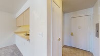 This single-floor residential unit offers a straightforward and functional layout designed for comfortable everyday living. The open living area is finished with plush wall-to-wall carpeting and painted in crisp, neutral white tones, creating a bright and welcoming atmosphere. A pass-through kitchen connects seamlessly to the living space, featuring light wood cabinetry, white laminate countertops, a coil-burner electric range with overhead ventilation, and a refrigerator — providing ample storage and prep space. The kitchen's open design allows for easy interaction between cooking and living areas. A carpeted hallway leads to the bedroom, which benefits from natural light through a large window with exterior views of greenery. The room also includes a wall-mounted HVAC unit for individualized climate control. The bathroom is finished with warm tile-style flooring and includes a light wood vanity with an integrated sink, a mirror, and accessibility grab bars — making it both practical and inclusive. Throughout the unit, white walls and consistent trim work lend a cohesive, clean aesthetic. The entry area, accessed via a gold-hardware door, transitions smoothly into the main living spaces, rounding out a well-organized and move-in-ready home.