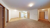 This single-floor unit presents an open-concept layout that seamlessly integrates the living area and kitchen into one cohesive space. The living area is finished with neutral-toned carpeting, providing a comfortable and warm underfoot feel, while the kitchen zone transitions to easy-maintenance hardwood-style flooring. The kitchen is equipped with white appliances, including a refrigerator and oven range, complemented by light wood cabinetry and a single-basin sink beneath under-cabinet fluorescent lighting. Two doors off the kitchen likely lead to additional utility or storage spaces. The walls throughout are painted in a clean, neutral white, enhancing the sense of openness. Flush-mount ceiling light fixtures provide consistent overhead illumination across the space. A textured ceiling adds subtle dimension overhead, while light wood baseboards frame each room with a cohesive trim detail. A thermostat and electrical outlets are conveniently positioned along the main wall of the living area. Overall, the space offers a straightforward, functional layout suited for everyday living.