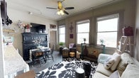 This single-floor space offers a warm and eclectic living environment filled with personality and charm. The main living area is anchored by a bold black-and-white floral area rug that draws the eye and sets the tone for the room's layered, decorative style. A ceiling fan with a light fixture overhead provides both comfort and ambient lighting, complementing the generous natural light that streams in through multiple large windows along the exterior wall. These windows also offer outward views, adding a sense of openness to the space.

The room seamlessly integrates a dining area, furnished with a round dark table and matching Windsor-style chairs, positioned beside a substantial dark-painted hutch that serves as a striking focal point. Decorative wall hangings, including a cross motif, add a personal and curated touch to the neutral-toned walls. Vintage-style trunks, decorative vases, and assorted accent pieces are thoughtfully arranged throughout, contributing to the space's layered aesthetic.

The concrete-style flooring grounds the room with an industrial sensibility, balancing the warmer decorative elements. Overall, the space presents a characterful and well-appointed interior with a distinctive, lived-in charm.
