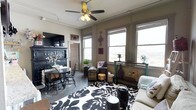 This single-floor space offers a warm and eclectic living environment filled with personality and charm. The main living area is anchored by a bold black-and-white floral area rug that draws the eye and sets the tone for the room's layered, decorative style. A ceiling fan with a light fixture overhead provides both comfort and ambient lighting, complementing the generous natural light that streams in through multiple large windows along the exterior wall. These windows also offer outward views, adding a sense of openness to the space.

The room seamlessly integrates a dining area, furnished with a round dark table and matching Windsor-style chairs, positioned beside a substantial dark-painted hutch that serves as a striking focal point. Decorative wall hangings, including a cross motif, add a personal and curated touch to the neutral-toned walls. Vintage-style trunks, decorative vases, and assorted accent pieces are thoughtfully arranged throughout, contributing to the space's layered aesthetic.

The concrete-style flooring grounds the room with an industrial sensibility, balancing the warmer decorative elements. Overall, the space presents a characterful and well-appointed interior with a distinctive, lived-in charm.
