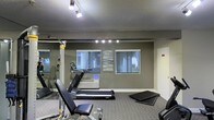 This single-floor fitness room is designed to provide a functional and focused workout environment. The space is outfitted with a selection of exercise equipment, including a treadmill for cardio training, a multi-use cable weight machine, an adjustable workout bench, and exercise mats for stretching or floor work — offering a well-rounded range of training options. The room features a textured ceiling with track lighting that illuminates the space effectively, supplemented by a row of accent lights mounted along one wall. The walls are finished in a neutral dark gray tone, lending the room a modern, gym-like atmosphere. Two windows along the back wall allow natural light to filter in and provide views of the surrounding greenery, creating a more open feel. A large wall mirror on one side enhances the sense of space and allows users to monitor their form during workouts. The flooring consists of low-pile carpet suited for the exercise setting. A wall-mounted unit beneath the windows suggests climate control for year-round comfort. Overall, this fitness room is a practical and well-appointed space for maintaining an active lifestyle.