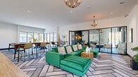 Located at 1000 Treesort View in Gainesville, GA, this single-floor community clubhouse delivers a sophisticated, resort-inspired atmosphere through bold design choices and thoughtfully curated spaces. The open-plan main lounge is anchored by a striking emerald green sectional sofa, set atop a large geometric herringbone-patterned area rug. Dramatic branch-style chandeliers hang from high ceilings, casting a warm glow over the space, while large floor-to-ceiling glass doors and windows flood the interior with abundant natural light and provide views of the surrounding landscape. A dedicated game room features a shuffleboard table, a pool table, and high-top communal tables with bar-height seating, all framed by expansive windows overlooking the outdoors. A private lounge nook, finished with richly textured teal-green wallcovering, offers an intimate setting with a marble-top table and curated seating for smaller gatherings or meetings. Throughout the space, curated artwork and lush greenery add warmth and character. Light hardwood flooring transitions seamlessly between zones, complementing the clean white walls adorned with large-scale contemporary art pieces. This well-appointed clubhouse provides a dynamic, welcoming environment ideal for socializing, working, and recreation.