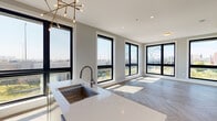This contemporary 2-bedroom, 2-bathroom residence in Providence, Rhode Island, offers a thoughtfully designed open-concept layout filled with natural light. The living and dining area is anchored by striking herringbone-patterned flooring and an expansive bank of black-framed floor-to-ceiling windows that frame captivating views of a nearby arched bridge and the surrounding cityscape. A sculptural sputnik chandelier adds a stylish focal point overhead.

The kitchen is outfitted with two-tone cabinetry in white and charcoal, a quartz waterfall island with an integrated sink, and stainless steel appliances including a range, microwave, and refrigerator — combining form and function seamlessly.

Both bedrooms continue the herringbone flooring and feature large black-framed windows that invite generous natural light and outdoor views. Each room includes individual climate control via wall-mounted mini-split units, ensuring year-round comfort.

The two bathrooms are finished in a clean, modern style. One features a floating wood-toned vanity with gold fixtures and a backlit mirror, complemented by large-format tile flooring and a glass-enclosed walk-in shower with crisp white tile surround. The overall palette of soft grays, white, and warm wood tones creates a cohesive and contemporary ambiance throughout.