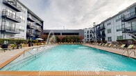 Located at 823 S Church St in Greenville, South Carolina, this generously sized outdoor amenity courtyard offers a resort-style living experience nestled within a modern multi-story residential complex. The centerpiece of the space is a large, crystal-clear swimming pool equipped with decorative water jet fountains, framed by wide tiled pool surrounds and bordered by warm-toned concrete decking that extends throughout the courtyard.

Ample sunbathing areas line both sides of the pool, furnished with green-cushioned lounge chairs that invite relaxation. Shaded seating clusters with umbrellas and green dining chairs provide comfortable outdoor gathering spots. A built-in outdoor kitchen station featuring multiple grills and stone-clad cabinetry serves as a dedicated space for outdoor entertaining.

Adding a playful touch, an oversized chess board with large game pieces is permanently installed on the courtyard floor, offering a unique social amenity. Raised landscaped planters with mature shrubs, ornamental trees, and boulders create natural green dividers throughout the space, softening the modern architectural backdrop.

The surrounding multi-story buildings feature bold blue and white facades with private balconies that overlook the courtyard, creating an enclosed, community-focused atmosphere. The space is expansive, designed to accommodate a wide range of outdoor leisure activities year-round.