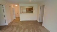 This single-floor residential space offers a straightforward, functional layout centered around an open living area with neutral-toned carpeting throughout. The focal point of the main room is a pass-through kitchen window, which connects the living space to the kitchen without fully separating the two areas. The kitchen features warm-toned wooden cabinetry, a white refrigerator, and light countertops, with recessed overhead lighting providing a well-lit workspace. The pass-through design enhances the sense of openness, allowing for easy interaction between the kitchen and living areas.

The living area is generously sized, providing ample room for furniture arrangement, and is finished with wall-to-wall carpeting in a neutral beige tone. Recessed ceiling lighting in the living space ensures consistent illumination throughout. Two doorways are visible from the main room — one leading to what appears to be an entryway or hallway with an exterior door, and another providing access to an adjacent room or storage area. The overall color palette is light and neutral, creating a clean, understated ambiance throughout the space.
