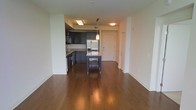 This single-floor space showcases a contemporary open-concept design that seamlessly connects the kitchen and living areas. Rich, dark hardwood flooring runs throughout, adding warmth and a polished aesthetic to the interior. The kitchen is a standout feature, outfitted with sleek dark cabinetry, granite or stone countertops, and stainless steel appliances including a French door refrigerator. A central kitchen island provides additional prep space and casual seating, while a pendant light overhead adds a modern touch. Recessed lighting illuminates the kitchen area, complementing the clean, neutral wall tones that keep the space feeling bright and open. The open layout offers a generous living area that flows naturally from the kitchen, making the space ideal for both everyday living and entertaining. A private entryway door is visible, providing a defined entry point to the unit. The overall ambiance is modern and well-appointed, with thoughtful design details that balance functionality and style throughout.