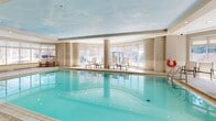 Located at 50 Laurier Ave E in Ottawa, this generously sized indoor aquatic and wellness facility offers a comprehensive suite of amenities across a single, open floor. The centrepiece is a large indoor swimming pool with turquoise-tiled water, framed by smooth round columns and surrounded by light beige ceramic tile flooring that extends throughout the space. Floor-to-ceiling windows line the pool area, flooding the interior with abundant natural light while offering views of the surrounding urban neighbourhood.

Poolside lounge chairs are arranged along the windowed perimeter, providing a comfortable retreat for relaxation. Adjacent to the pool, a dedicated fitness centre is visible through glass partitions, housing a variety of exercise equipment to support a full workout routine. A warm, wood-panelled sauna and steam room area is tucked to one side, featuring classic timber-clad walls, a glass-door steam room, and benches for a traditional spa experience.

The ceiling above the pool area is finished with a decorative sky-motif mural, adding a distinctive and airy character to the space. The overall ambiance is clean, bright, and resort-like, offering residents a refined wellness environment within the heart of Ottawa's downtown core.