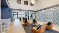 Located at 7570 32nd St N in Oakdale, Minnesota, this thoughtfully designed single-floor amenity space offers a generous footprint across a single level. The grand entrance lobby immediately impresses with soaring double-height ceilings, abundant natural light streaming through oversized windows, and light-toned hardwood-style flooring. A bold navy and white color palette ties the space together, accented by a striking geometric patterned feature wall. The central lounge area is anchored by a grouping of cognac leather swivel chairs arranged around a terrazzo coffee table beneath an elegant pendant light fixture. A separate lounge features a dramatic floor-to-ceiling stone fireplace flanked by flat-screen TVs and a grid-patterned navy accent wall, creating a warm and sophisticated gathering space. The kitchenette is well-appointed with marble-look countertops, white cabinetry, stainless steel appliances, and a matte black faucet. Multiple dining areas offer versatile seating options, including intimate upholstered banquette nooks with patterned wallpaper accents and pendant lighting. A private workspace with a natural wood desk and outdoor views completes the offering. Hallways connect the various zones seamlessly, enhanced by greenery and curated décor throughout.