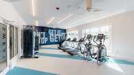 This well-appointed fitness center in Panama City Beach, Florida, offers a comprehensive workout experience within a single-floor layout designed with both functionality and style in mind. The main exercise area is anchored by a row of treadmills, ellipticals, and stair climbers positioned along large windows that flood the space with natural light and provide views of the surrounding palm-lined outdoor amenities. A bold, wave-patterned motivational mural in deep navy blue serves as a striking focal point, reinforcing the coastal character of the facility. The flooring features a distinctive multi-tone stripe design in teal, white, and green, adding visual energy throughout. A dedicated strength training zone offers a functional trainer cable machine, weight benches, and a full dumbbell rack, while a wall-mounted television and built-in white cabinetry with marble-patterned shelving provide both utility and a clean, modern aesthetic. Modern ceiling fans with wood-toned blades complement the linear LED lighting overhead. A separate studio room, accessible through a frosted glass door labeled "Stretch," is equipped with a spin bike, stability balls, BOSU trainers, plyo boxes, and a large mirrored wall beneath an artistic navy mural depicting yoga poses—ideal for group classes or individual training sessions.