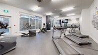 Located at 1452 Brenton Street in Halifax, NS, this fitness center offers a single, thoughtfully arranged exercise room that provides a comprehensive workout environment. The space features durable rubber flooring throughout, bright recessed and fluorescent panel lighting, and crisp white walls accented by a bold fitness-themed typographic mural that energizes the atmosphere.

Large windows line the perimeter, flooding the room with natural light and offering views of the surrounding urban streetscape. A full-length mirror along one wall enhances the sense of openness and allows users to monitor their form during workouts. A wall-mounted flat-screen TV adds a convenient entertainment option for cardio sessions.

The cardio zone is well-stocked with multiple treadmills, elliptical trainers, a stair climber, and stationary and spin bikes. The strength training area includes a set of adjustable benches, dual dumbbell racks with a full range of weights, and medicine balls, providing everything needed for a complete resistance workout. A dark accent column serves as a subtle architectural divider between zones, adding visual interest without disrupting the open layout.

Ventilation is managed through ceiling-mounted HVAC units, ensuring a comfortable environment year-round. The facility is clean, modern, and efficiently organized to maximize usable floor space for a variety of fitness activities.
