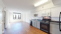 This single-floor unit presents a clean, modern aesthetic throughout, characterized by crisp white walls, smooth ceilings, and warm hardwood-style flooring that flows consistently across the main living areas. The open layout connects the living space directly to the kitchen, creating a spacious, airy feel enhanced by an abundance of natural light streaming through large black-framed windows that frame views of the surrounding urban streetscape.

The kitchen is well-appointed with white upper cabinetry, gray lower cabinets, dark granite-style countertops, and a mosaic tile backsplash that adds a subtle decorative touch. Black appliances — including a gas range, dishwasher, built-in microwave, and full-size refrigerator — complete the functional setup.

The living area is generously proportioned, with multiple windows allowing natural light to illuminate the space throughout the day. Additional rooms, visible in the floor plan, provide flexible living arrangements, each featuring the same consistent flooring and neutral wall tones.

The bathroom offers white tile flooring, a vanity with gray cabinetry, a large mirror with overhead lighting, and accessibility-friendly grab bars, rounding out this well-designed, light-filled unit.