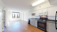 This single-floor unit presents a clean, modern aesthetic throughout, characterized by crisp white walls, smooth ceilings, and warm hardwood-style flooring that flows consistently across the main living areas. The open layout connects the living space directly to the kitchen, creating a spacious, airy feel enhanced by an abundance of natural light streaming through large black-framed windows that frame views of the surrounding urban streetscape.

The kitchen is well-appointed with white upper cabinetry, gray lower cabinets, dark granite-style countertops, and a mosaic tile backsplash that adds a subtle decorative touch. Black appliances — including a gas range, dishwasher, built-in microwave, and full-size refrigerator — complete the functional setup.

The living area is generously proportioned, with multiple windows allowing natural light to illuminate the space throughout the day. Additional rooms, visible in the floor plan, provide flexible living arrangements, each featuring the same consistent flooring and neutral wall tones.

The bathroom offers white tile flooring, a vanity with gray cabinetry, a large mirror with overhead lighting, and accessibility-friendly grab bars, rounding out this well-designed, light-filled unit.