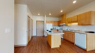 This single-floor residential unit offers a thoughtfully designed layout that balances functionality and comfort. The kitchen serves as a central highlight, featuring warm honey-toned cabinetry, granite-look countertops, a central island with breakfast bar seating, and a full suite of white appliances including a refrigerator, electric range, microwave, and dishwasher. Recessed lighting and rich hardwood-style flooring give the kitchen and adjacent living area a cohesive, inviting feel. The living area is open and airy, benefiting from a large window that draws in generous natural light and offers views of the surrounding greenery. Neutral walls and clean lines throughout create a versatile backdrop adaptable to a variety of furnishing styles. The bedrooms are carpeted for comfort and feature large windows fitted with blinds, allowing for excellent light control while maintaining a connection to the outdoors. The bathroom is finished with slate-style tile flooring, a warm wood vanity, a wide mirror, and grab bars, indicating accessible design considerations. The unit's overall aesthetic is clean and modern, with consistent use of warm wood tones, neutral palettes, and practical finishes throughout.