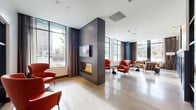 Located at 260 Portland Ave in Minneapolis, MN, this thoughtfully designed ground-floor lobby and amenity space offers a generous and well-appointed environment for residents and visitors alike. The interior is anchored by a striking dark-paneled, double-sided fireplace with a mounted display above, serving as a dramatic focal point that divides the main lounge into two distinct seating zones. One side features bold red accent chairs arranged around low side tables, while the other showcases a mix of blue and red mid-century-style seating bathed in natural light from floor-to-ceiling windows overlooking Portland Avenue. Polished concrete floors run throughout, lending an industrial-modern aesthetic that is complemented by warm wood ceiling accents and dark navy architectural columns. A communal dining or coworking area features a richly textured table with red shell chairs beneath a sculptural pendant light fixture, accompanied by original wall art. Glass-partitioned rooms provide additional semi-private workspaces. The mail area includes a full bank of individual mailboxes, a water fountain station, and direct access to a fitness center visible through interior windows. Package concierge lockers and stainless steel elevators are conveniently located near the building entrance, completing this fully functional and design-forward lobby experience.