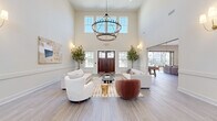 This generously proportioned community living space is designed to impress from the moment you enter through its rich, dark-wood double doors. The grand foyer opens into a dramatic main lounge area defined by soaring double-height ceilings and clerestory windows that flood the interior with abundant natural light. Light gray wood-look flooring flows throughout, complementing the crisp white walls and classic wainscoting detail.

The main seating area is anchored by a striking two-tiered iron chandelier and features a curated arrangement of plush white sofas and accent chairs, paired with a patterned area rug and potted olive trees that bring a touch of organic warmth. Adjacent to the lounge, a dedicated entertainment zone offers a wall-mounted flat-screen TV, comfortable seating, and a shuffleboard table, perfect for social gatherings.

A wet bar with counter seating provides an ideal spot for casual entertaining, while a stylish beverage and refreshment station with decorative shelving and a framed mirror adds a refined touch. The space also includes a quiet nook with a desk for focused work or meetings.

Large sliding glass doors open directly to an outdoor terrace overlooking a resort-style pool, creating a seamless indoor-outdoor living experience bathed in natural light.