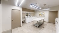 Located at 2220 Weston Road in Toronto, this shared laundry facility is a well-organized and efficiently designed space that serves its practical purpose with a clean, modern aesthetic. The room is finished with large-format, light-coloured floor tiles that contribute to a bright and hygienic atmosphere, complemented by beige ceramic wall tiles that line the machine area and add a subtle warmth to the interior.

The facility is equipped with a row of commercial-grade front-load washers and dryers, each outfitted with digital control panels for ease of use. A deep utility sink is also available for hand-washing needs. Central to the room is a spacious, white quartz-topped folding table with a stainless steel frame, providing generous surface area for sorting and folding laundry. An additional stainless steel utility table offers further workspace.

Overhead, a combination of recessed ceiling lights and modern suspended linear LED fixtures ensures the entire room is evenly and brightly lit. A framed bulletin board mounted on the wall displays informational notices for users. A grey upholstered bench along one wall provides a comfortable waiting area. The space is accessed via a solid door with a frosted glass panel, and ventilation is managed through ceiling-mounted HVAC registers, maintaining a comfortable environment throughout.
