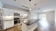 Located at 128 Mill St in Clayton, NC, this single-story home offers five bedrooms and two bathrooms across a well-designed, single-level floor plan. The open-concept kitchen and living area serves as the heart of the home, showcasing crisp white cabinetry, a white subway tile backsplash, stainless steel appliances, and a large center island with a dark granite countertop. Sleek pendant lights and track lighting illuminate the space, while rich hardwood-style flooring flows seamlessly into the living area, which features a ceiling fan and French doors that invite natural light and outdoor access. Both bathrooms are finished with white vanity cabinetry, dark granite countertops, large mirrors, and hardwood-style flooring. The primary bathroom includes a glass-enclosed shower and direct access to a walk-in closet with wire shelving. The bedrooms are carpeted and bright, with multiple windows fitted with blinds and ceiling fans for comfort. Additional bedrooms also include closets with wire shelving systems. A dedicated laundry room with washer and dryer hookups adds everyday convenience. Two hallways connect the rooms efficiently throughout the layout, completing this functional and modern home.