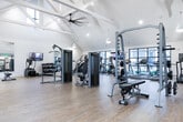 Located at 1100 Enterprise Way NW in Huntsville, Alabama, this single-floor fitness center is designed to support a full range of workout needs in a bright, modern setting. The facility is characterized by soaring vaulted ceilings with exposed white beam detailing, pendant lighting, and an abundance of natural light streaming through large, black-framed windows that offer views of the surrounding community grounds and pool area.

The main workout floor features light wood-look flooring and is outfitted with an extensive array of strength training equipment, including cable machines, weight benches, and functional training stations with medicine balls and resistance tools. A wall-mounted television and large mirrors enhance the training environment throughout.

A dedicated cardio zone houses multiple treadmills, ellipticals, stair climbers, and rowing machines, anchored by hexagonal tile flooring near the main entrance. A separate cycling studio features Echelon smart bikes and additional stationary cycles positioned before a sweeping floor-to-ceiling window wall, with a full-length mirror wall completing the studio setup. A barre and stretching area further rounds out the facility's offerings.

The entry area includes a clean, paneled white accent wall with artwork, creating a welcoming transition into the fitness space. Throughout, the design balances functionality with a polished, contemporary aesthetic.