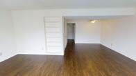 This two-floor space combines practical design with classic details throughout. The main living area is highlighted by rich, dark hardwood floors that extend seamlessly through the open layout, creating a warm and grounded atmosphere. A built-in shelving unit along one wall provides ample display and storage space, adding both function and architectural character to the room. A ceiling fan overhead ensures year-round comfort while complementing the clean, understated aesthetic of the space. White walls throughout keep the interior feeling bright and open, allowing natural light to reflect evenly across the rooms. A hallway leads further into the layout, connecting the living areas to additional rooms and offering a sense of depth and flow within the floor plan. The open passage and well-proportioned spaces make the layout easy to navigate and highly adaptable for a variety of uses. The second floor expands the overall footprint of the property, providing additional living or functional areas beyond the main level. Overall, the space presents a straightforward and well-maintained interior, characterized by consistent finishes, a neutral color palette, and thoughtful built-in features that enhance everyday livability.