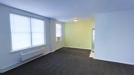 This single-floor space offers a clean and functional layout designed for comfortable everyday living. The main living area is finished with dark wall-to-wall carpeting that contrasts neatly against light-colored walls, with one accent wall painted in a soft yellow tone that adds warmth to the room. Two generously sized windows fitted with white horizontal blinds allow ample natural light to fill the space, while a window-mounted air conditioning unit provides climate control. A baseboard heating unit runs along the exterior wall, ensuring year-round comfort. Flush-mount ceiling lighting provides even illumination throughout the room. The living area opens toward an adjacent space, visible in the background, which features hard flooring and appears to connect to a kitchen area, suggested by the presence of a refrigerator. The overall layout is open and uncluttered, offering flexibility for a variety of furniture arrangements. The neutral tones, practical finishes, and straightforward design make this a versatile and move-in ready space suited for a range of occupants.
