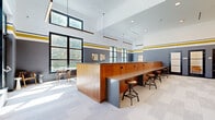 Located at 201 Munson Street in New Haven, CT, this thoughtfully designed single-floor space offers a dynamic and functional environment ideal for collaboration and gatherings. The interior is defined by soaring ceilings that create an open, airy atmosphere, complemented by large black-framed windows along multiple walls that flood the space with natural light and provide views of the surrounding greenery.

The design palette centers on slate gray walls accented by bold gold and white horizontal striping near the ceiling line, lending the space a contemporary yet spirited character. Light-toned carpet tiles cover the floors throughout, adding warmth and acoustic comfort.

A prominent warm-wood counter runs along one side of the room, lined with industrial-style adjustable bar stools, serving as an ideal workspace or casual gathering bar. Opposite, floor-to-ceiling windows frame a sunlit seating area furnished with sleek tables and wooden-framed chairs. A cozy booth nook features tufted black leather banquette seating paired with minimalist black pedestal tables and decorative gold sconce lighting.

Along one wall, a row of private rooms enclosed by black-framed glass doors provides quieter, semi-enclosed spaces for focused work or small meetings. Curated artwork and thoughtful lighting fixtures throughout complete this well-appointed, multi-functional lounge environment.