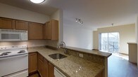 This single-floor space showcases a thoughtfully designed open-concept layout that seamlessly connects the kitchen to the living area. The kitchen is equipped with warm-toned wood cabinetry providing ample storage, and features polished granite countertops with a distinctive speckled pattern in neutral tones. A stainless steel sink with a modern arc-neck faucet is centrally positioned along the counter, complementing the clean, contemporary aesthetic. A track lighting fixture overhead provides directed task lighting throughout the kitchen space.

The kitchen flows naturally into the adjacent living area, which benefits from a large sliding glass door or window that introduces generous natural light and provides access to what appears to be a balcony or outdoor space with a railing visible beyond. The living area features neutral-toned flooring and light-colored walls, creating a bright and airy ambiance throughout. The open sightlines between the kitchen and living area enhance the overall sense of space, making the layout feel connected and functional. The combination of natural light, quality finishes, and open design gives this space a welcoming and practical character.
