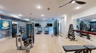 Located at 720 Verdae Blvd in Greenville, South Carolina, this fitness center offers a thoughtfully designed exercise space with a comfortable and motivating atmosphere. The room features light hardwood-style flooring throughout, recessed lighting, and multiple ceiling fans that ensure airflow and comfort during workouts.

The space is outfitted with a robust selection of Matrix-brand equipment, including treadmills, a cable machine, a leg press, an elliptical, a functional trainer, and a weight bench. A full dumbbell rack provides a range of free weights, while a large wall-mounted mirror amplifies the sense of space and supports proper form during exercise. Two flat-screen TVs are mounted on the wall near the cardio machines for entertainment during workouts.

A standout feature is the textured chevron-patterned accent wall in a cool blue-gray tone, complemented by motivational fitness artwork that energizes the environment. Natural light filters in through a window dressed with sheer curtains, adding brightness to the space. Built-in cabinetry with rich wood-toned finishes provides storage along one wall. The fitness center also includes a Matrix vertical training unit and a flat workout bench, rounding out the comprehensive range of equipment available to users.