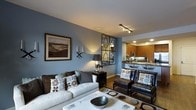 This well-appointed single-floor residence offers a thoughtfully designed open-concept layout that seamlessly connects the living area to the kitchen. The living room is anchored by cool slate-blue accent walls that create a calm, sophisticated atmosphere, complemented by warm hardwood flooring that runs throughout the space. A plush white sofa and a pair of dark leather armchairs provide ample seating, accented by decorative throw pillows that echo the room's blue and brown color palette. A framed landscape painting, wall sconces, and a striking geometric mirror-and-metal wall accent add visual interest and character to the space. Track lighting on the ceiling ensures the area is well-lit and functional. The kitchen, visible from the living room, features warm honey-toned wood cabinetry, a granite or stone-topped breakfast bar with seating, stainless steel appliances, and recessed lighting, making it both practical and inviting. The open sightlines between the kitchen and living room enhance the sense of spaciousness and encourage easy flow for everyday living and entertaining. Overall, the space exudes a modern yet cozy aesthetic, blending contemporary design elements with warm, inviting tones.