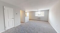 This single-floor residence offers a thoughtfully organized layout across several well-defined living spaces. The expansive living room is finished with soft wall-to-wall carpeting in a neutral gray tone, complemented by light gray walls that lend a clean, airy atmosphere. A ceiling fan overhead and a wall-mounted air conditioning unit ensure year-round comfort, while a large window invites an abundance of natural light and outdoor views of surrounding greenery. A half-wall partition separates the living area from the kitchen, creating an open yet defined flow between spaces. The kitchen is well-equipped with warm-toned wood cabinetry, a slate-blue tile backsplash, light countertops, and a full suite of stainless steel appliances including a gas range, microwave, and refrigerator. Tile flooring adds a practical, easy-to-maintain surface underfoot. Both bedrooms are carpeted and bathed in natural light from large windows, with one featuring a sliding double-door closet. The residence includes two bathrooms: one with a tub-shower combination accented by classic black-and-white tile detailing, and a second with a glass-door walk-in shower and sleek modern fixtures. The overall ambiance is bright, neutral, and move-in ready.