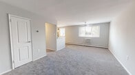 This single-floor residence offers a thoughtfully organized layout across several well-defined living spaces. The expansive living room is finished with soft wall-to-wall carpeting in a neutral gray tone, complemented by light gray walls that lend a clean, airy atmosphere. A ceiling fan overhead and a wall-mounted air conditioning unit ensure year-round comfort, while a large window invites an abundance of natural light and outdoor views of surrounding greenery. A half-wall partition separates the living area from the kitchen, creating an open yet defined flow between spaces. The kitchen is well-equipped with warm-toned wood cabinetry, a slate-blue tile backsplash, light countertops, and a full suite of stainless steel appliances including a gas range, microwave, and refrigerator. Tile flooring adds a practical, easy-to-maintain surface underfoot. Both bedrooms are carpeted and bathed in natural light from large windows, with one featuring a sliding double-door closet. The residence includes two bathrooms: one with a tub-shower combination accented by classic black-and-white tile detailing, and a second with a glass-door walk-in shower and sleek modern fixtures. The overall ambiance is bright, neutral, and move-in ready.