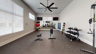 This fitness facility is thoughtfully designed to accommodate a wide range of workout styles and fitness goals. The space is organized across multiple interconnected areas, each dedicated to a specific type of training. Soaring ceilings throughout lend an open, airy feel, while speckled rubber flooring provides both durability and comfort underfoot.

The cardio area is outfitted with multiple treadmills, an elliptical trainer, a recumbent bike, and a wall-mounted flat-screen television, creating an engaging environment for sustained aerobic exercise. Natural light streams in through large windows fitted with blinds, brightening the space considerably. An exterior door provides direct outdoor access.

A dedicated strength training room features a full rack of dumbbells, medicine balls, weight benches, and a large wall mirror that visually expands the space. Dual wall-mounted screens and a ceiling fan with integrated lighting enhance both functionality and comfort. An adjoining area houses a cable crossover machine, ideal for versatile resistance training.

A separate cycling area accommodates two connected spin bikes positioned in front of oversized windows, offering a light-filled, focused setting for indoor cycling sessions. Together, these zones form a comprehensive, well-appointed fitness environment.