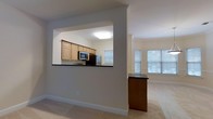 This two-floor space showcases a thoughtfully designed open-concept layout that seamlessly connects the kitchen and living areas. The kitchen is framed by a distinctive pass-through window cut into the wall, offering a view into the main living space while maintaining a sense of defined zones. Light wood cabinetry lines the kitchen walls, complemented by dark countertops that provide a striking contrast. Black appliances, including a built-in microwave and refrigerator, add a cohesive, modern touch. A raised breakfast bar with matching dark countertop extends from the kitchen's partition wall, creating a convenient casual dining or prep surface. The adjacent living area is generously sized, featuring neutral carpet flooring and soft, light grey walls that lend a clean, understated aesthetic throughout. Large windows on multiple walls flood the living space with abundant natural light, enhancing the open and airy feel of the interior. The overall ambiance is calm and functional, with a neutral palette that provides a versatile backdrop. The layout across two floors offers ample space for a variety of living arrangements.