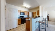 This single-floor residential unit offers a practical and comfortable layout designed for everyday living. The kitchen is a functional centerpiece, featuring warm wood cabinetry, dark countertops, black appliances including a range and over-the-range microwave, and durable wood-look flooring. A peninsula with a dark countertop and bar seating connects the kitchen to the adjacent living and dining areas, making it ideal for casual dining and entertaining.

The living room is bright and welcoming, with large windows that invite generous natural light and provide outdoor views. Plush carpeting, a ceiling fan, and access to a private exterior door enhance the comfort and functionality of the space.

The bedrooms are carpeted and well-proportioned, each offering windows with natural light. One bedroom includes a ceiling fan and ample space for furniture arrangement, while another features a window with blinds and space for a dedicated work area.

The bathroom is cleanly appointed with a wood-toned vanity, dark countertop, a large mirror, and a shower-tub combination. An in-unit laundry area provides added convenience, housing a top-load washer alongside built-in shelving overhead and wood-look flooring underfoot. The overall ambiance is neutral and move-in ready throughout.